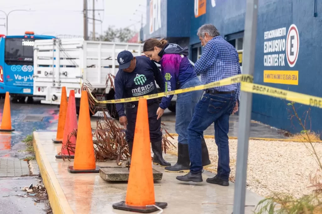 Personas con ropa de trabajo inspeccionando drenaje en banqueta mientras llueve, con conos de señalización y cinta amarilla