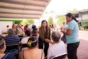 Mujer joven saludando de mano a una persona mayor sentada en un evento comunitario con más asistentes al fondo