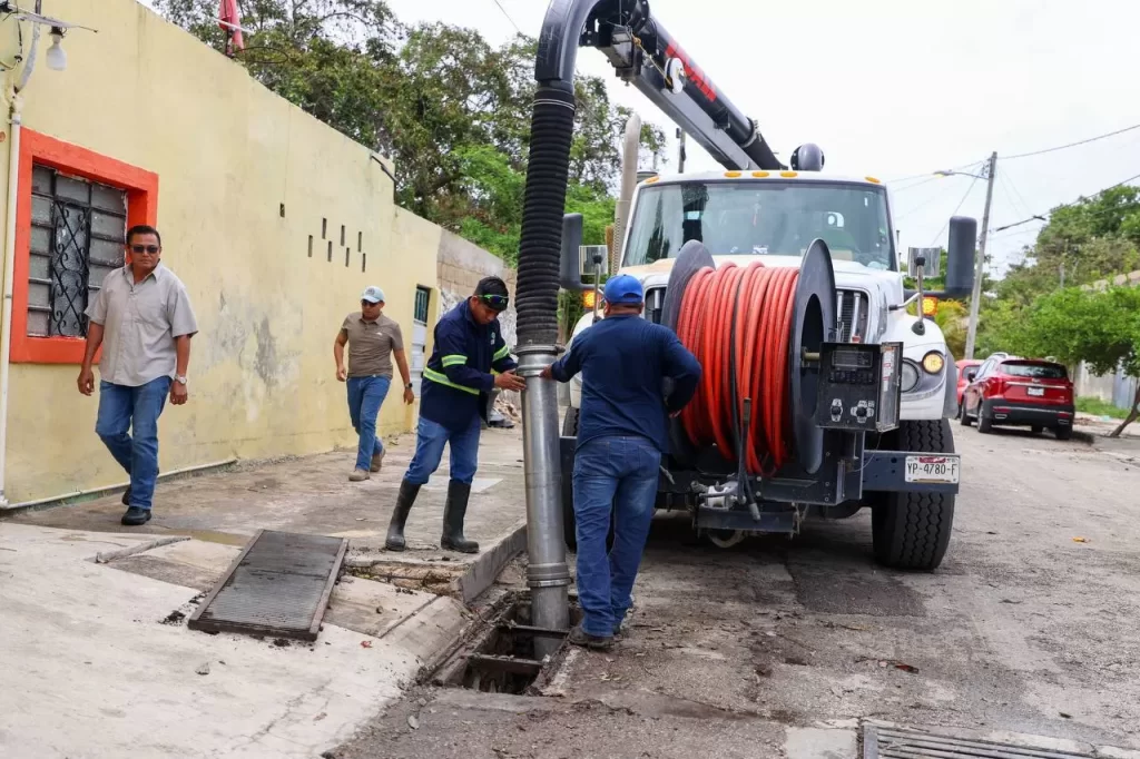Dos trabajadores operando equipo de limpieza de drenaje en una calle mientras dos hombres caminan cerca