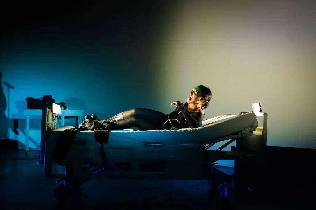 Mujer recostada en cama hospitalaria bajo luces dramáticas, escena teatral expresiva