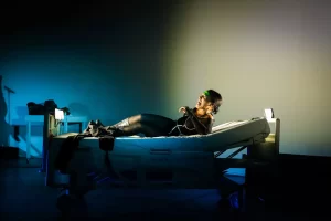 Mujer recostada en cama hospitalaria bajo luces dramáticas, escena teatral expresiva