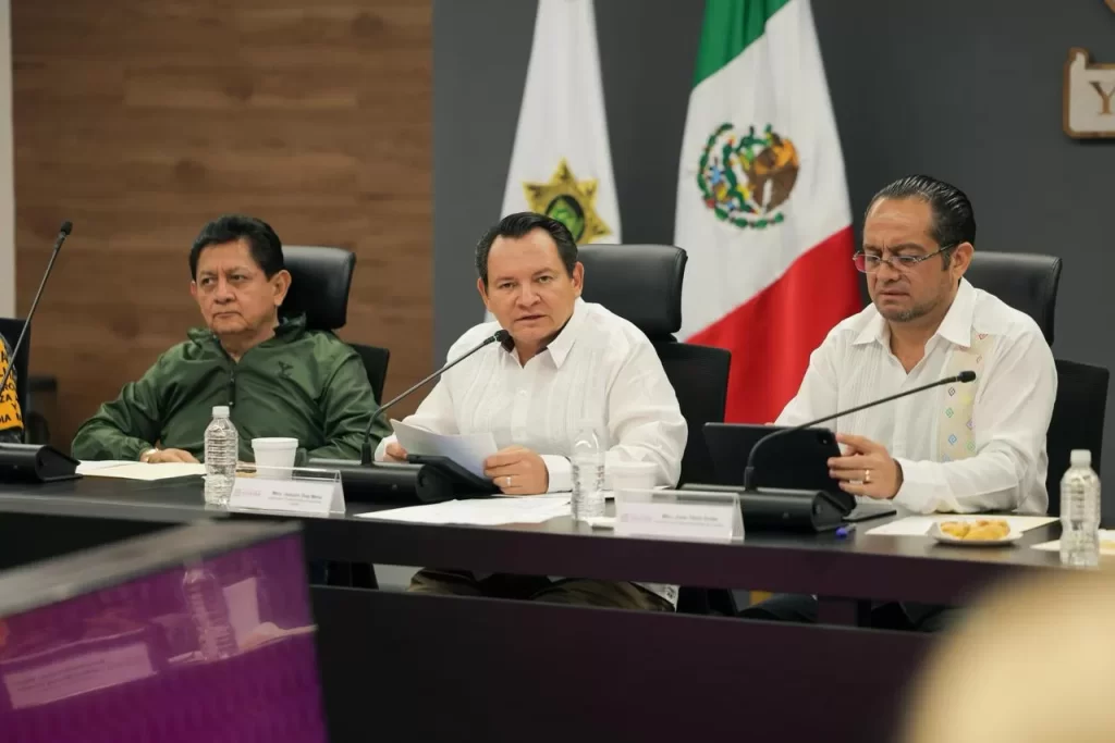 Tres hombres sentados en mesa con papeles y micrófonos, bandera de México y bandera institucional al fondo