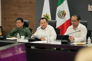 Tres hombres sentados en mesa con papeles y micrófonos, bandera de México y bandera institucional al fondo