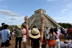 Turistas observando la Pirámide de Kukulkán en Chichén Itzá, con mujer de espaldas con sombrero levantando las manos