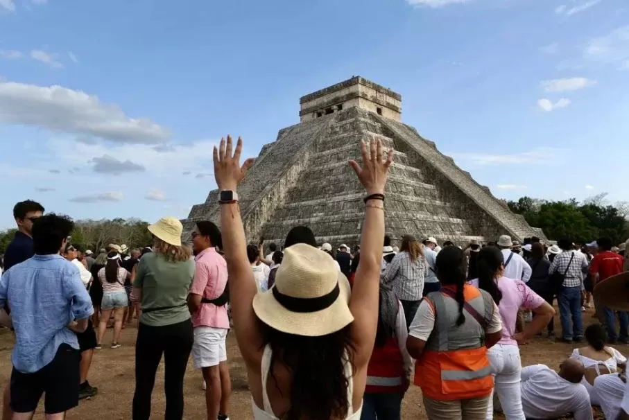 Turistas observando la Pirámide de Kukulkán en Chichén Itzá, con mujer de espaldas con sombrero levantando las manos
