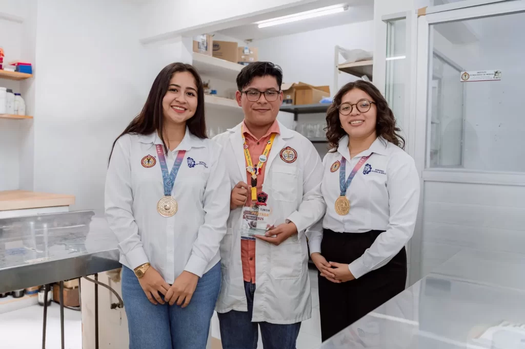 Tres jóvenes estudiantes en laboratorio usando batas blancas y medallas doradas, uno sostiene un trofeo