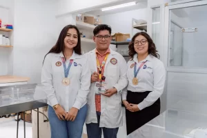 Tres jóvenes estudiantes en laboratorio usando batas blancas y medallas doradas, uno sostiene un trofeo