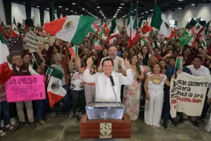 Hombre en podio con señoras y señores detrás con banderas mexicanas y pancartas en evento en Yucatán