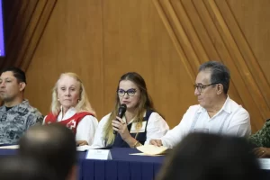 Mujer con gafas hablando al micrófono entre personas, una con uniforme militar, otra con chaleco rojo de Cruz Roja y un hombr