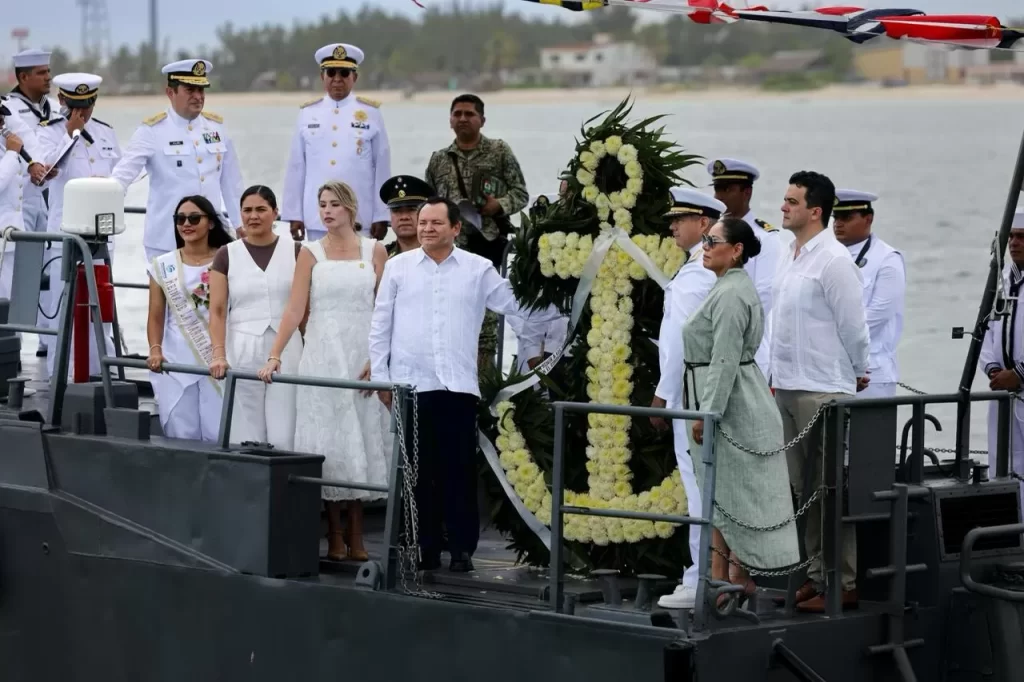 Personas en ceremonia naval sobre un bote con ofrenda floral en forma de ancla