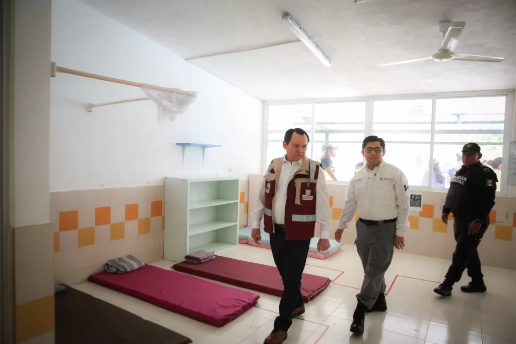 Dos hombres con chalecos oficiales y un policía dentro de una habitación con colchonetas en el piso y un ventilador