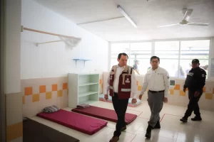 Dos hombres con chalecos oficiales y un policía dentro de una habitación con colchonetas en el piso y un ventilador