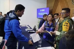 Personas en reunión del Consejo Municipal de Protección Civil de Mérida, una mujer con chaleco azul entrega documentos a un h