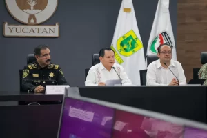 Tres hombres sentados en una mesa con micrófonos en una sala de conferencias, detrás banderas y escudo de Yucatán