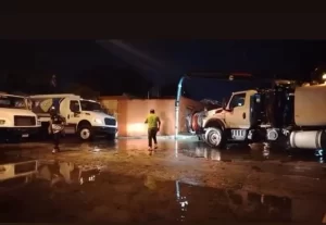 Dos camiones blancos estacionados y dos hombres caminando en terreno mojado durante la noche con luces encendidas