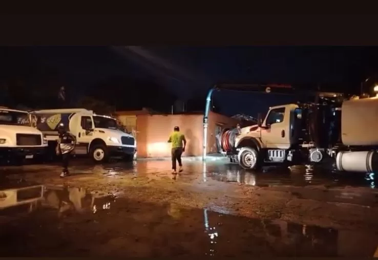 Dos camiones blancos estacionados y dos hombres caminando en terreno mojado durante la noche con luces encendidas