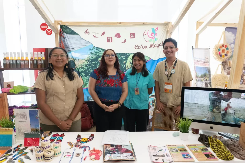 Cuatro personas sonrientes frente a un puesto con artesanías y un cartel de Co'ox Mayab