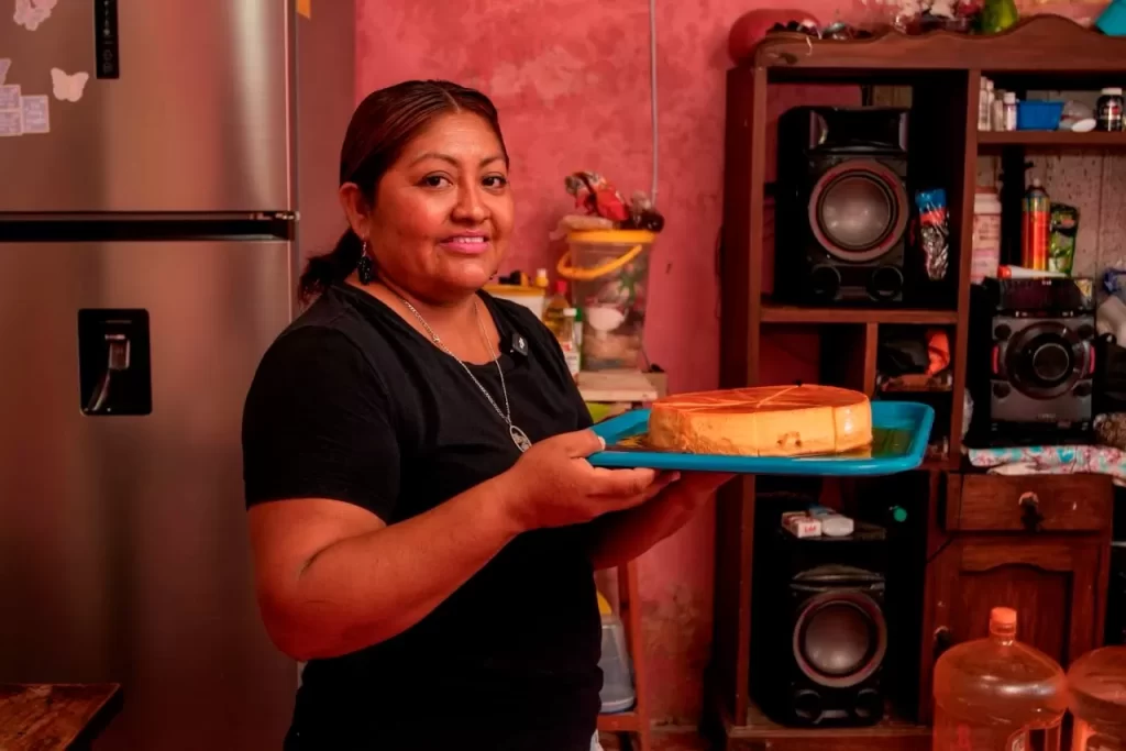 Mujer sonriente sostiene un pastel redondo en una bandeja azul en una cocina con paredes rosas