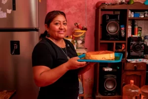 Mujer sonriente sostiene un pastel redondo en una bandeja azul en una cocina con paredes rosas