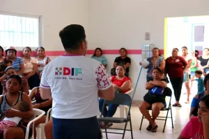 Hombre con camiseta del DIF Yucatán 2024-2030 hablando ante un grupo de personas sentadas y de pie en un salón interior