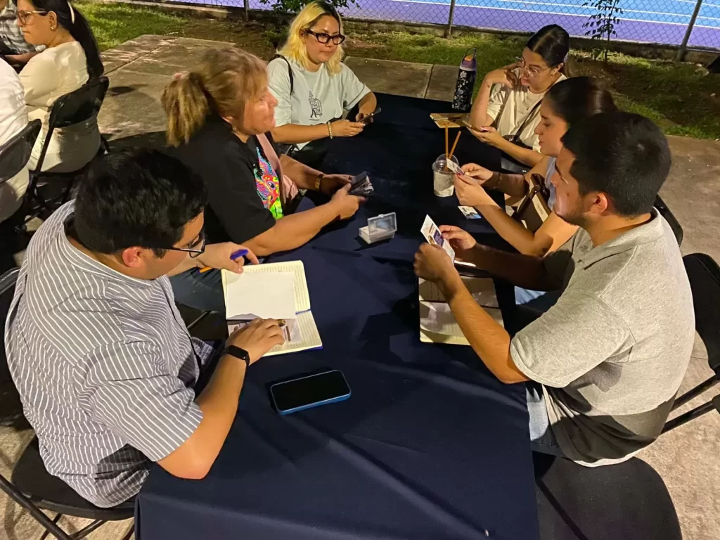 Seis personas reunidas en una mesa con mantel azul, algunas revisan tarjetas y anotan en libretas