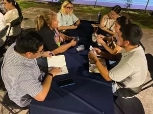 Seis personas reunidas en una mesa con mantel azul, algunas revisan tarjetas y anotan en libretas