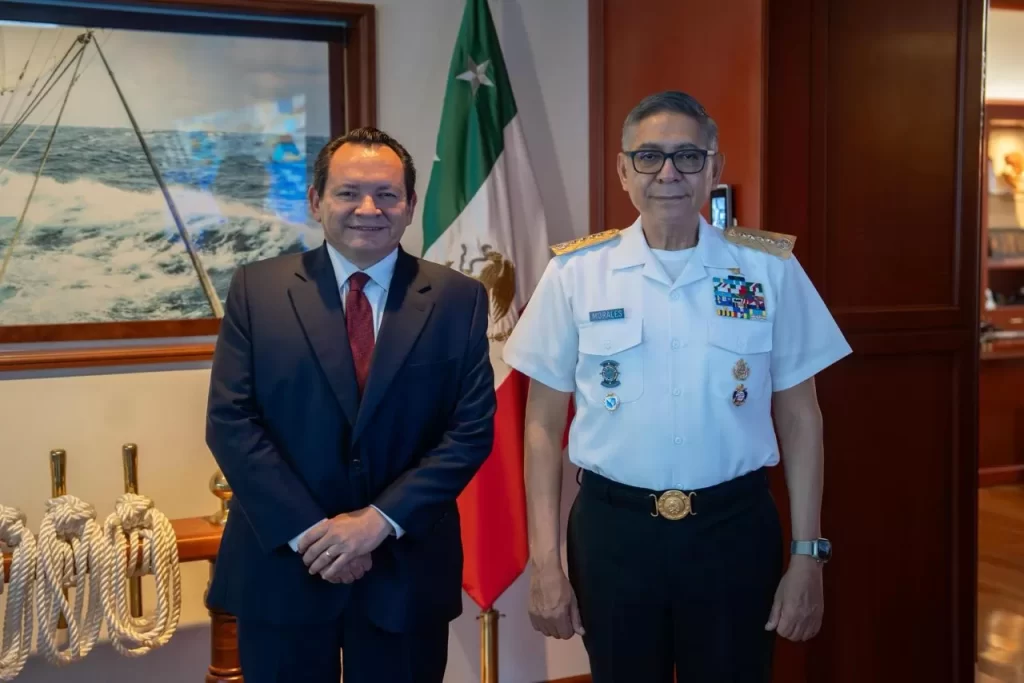 Dos hombres posando, uno con traje formal y otro con uniforme naval, junto a bandera de México