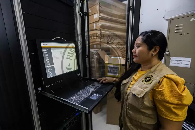 Mujer con chaleco de Fiscalía frente a una computadora en una oficina con cajas al fondo