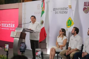 Hombre dando discurso en podio con bandera de México y bandera estatal de Yucatán, personas sentadas al lado