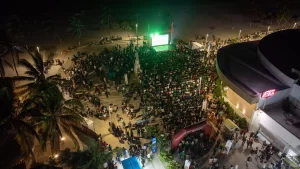 Personas reunidas al aire libre frente a una pantalla gigante en la noche, en una zona con palmeras y un edificio con letrero