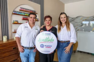 Tres personas sonrientes sostienen cartel de acreditación Mi Primer Crédito en espacio interior