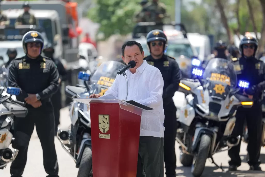 Hombre habla en podio con escudo de Yucatán rodeado por policías estatales con motos y camionetas