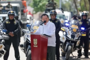 Hombre habla en podio con escudo de Yucatán rodeado por policías estatales con motos y camionetas