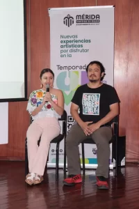 Dos personas sentadas frente a un cartel promocional de experiencias artísticas en Mérida, ella con blusa bordada y él con ca