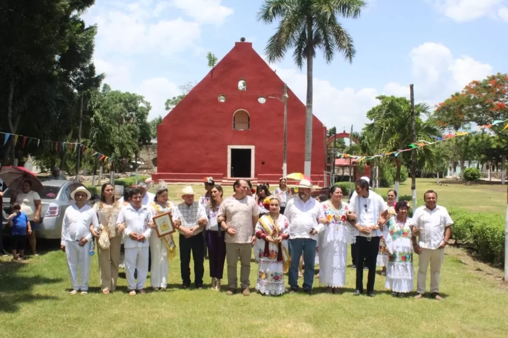 Personas vestidas con ropa tradicional mexicana paradas en fila frente a una iglesia de fachada roja