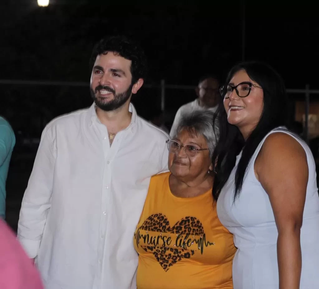 Hombre con camisa blanca, mujer mayor con camiseta amarilla y mujer con vestido blanco posan sonriendo