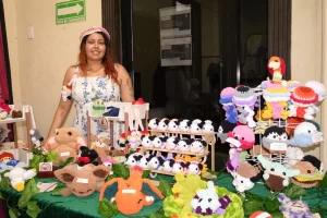 Mujer con vestido blanco de flores azules y gorro rosa detrás de una mesa con juguetes tejidos a mano de varios colores y for