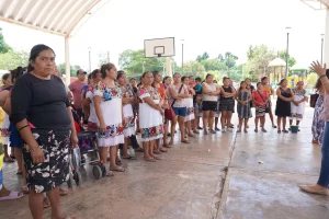 Grupo de mujeres y algunos hombres en una reunión bajo una cancha techada de básquetbol, algunas mujeres con vestidos tradici