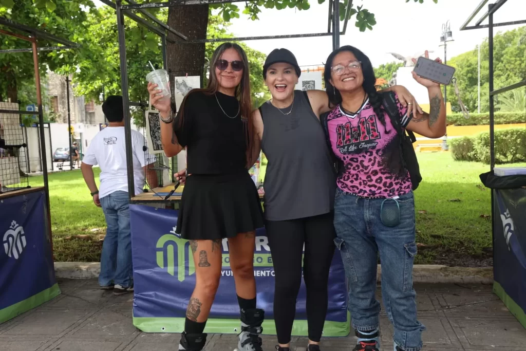 Tres mujeres jóvenes posando sonrientes al aire libre, una con falda negra, otra con camiseta gris y gorra, y otra con camise