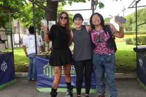 Tres mujeres jóvenes posando sonrientes al aire libre, una con falda negra, otra con camiseta gris y gorra, y otra con camise