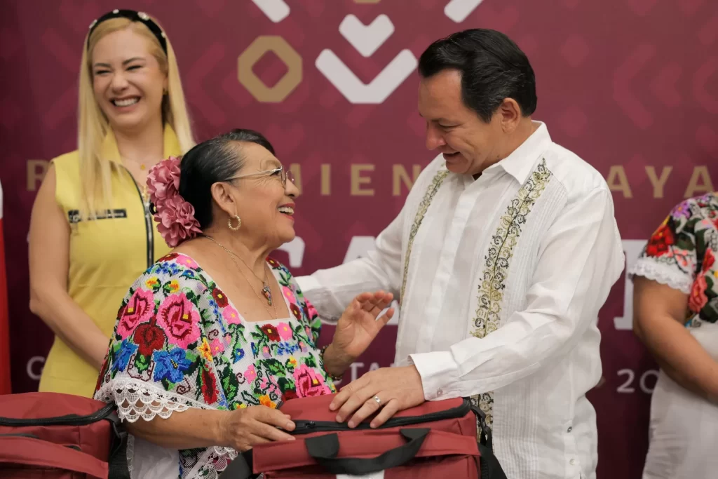 Hombre vestido de blanco entrega mochila a mujer con blusa tradicional y flores en el cabello
