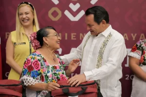 Hombre vestido de blanco entrega mochila a mujer con blusa tradicional y flores en el cabello