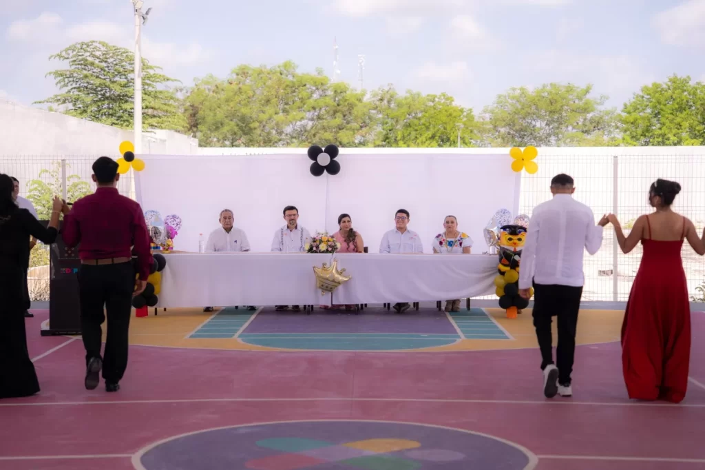 Personas formadas en parejas caminando hacia mesa con cinco personas sentadas decorada con globos en gimnasio escolar