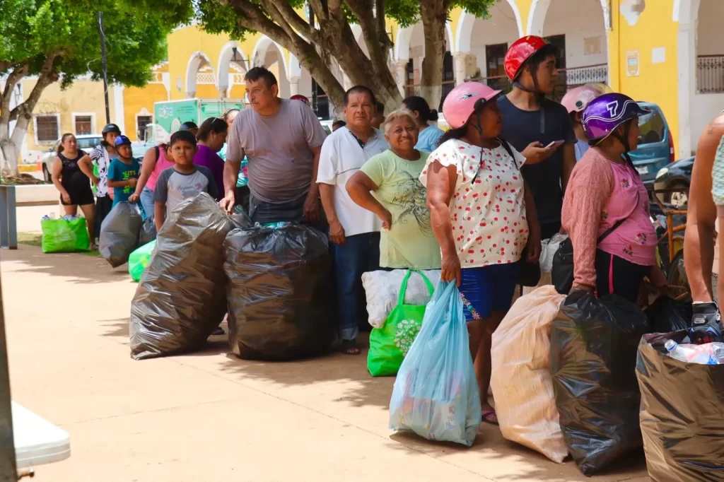 Grupo de personas en fila sosteniendo bolsas grandes de basura y de plástico, algunas con casco, bajo árboles