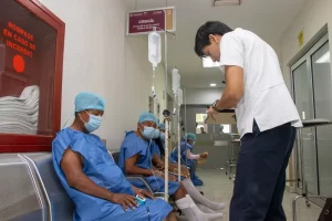 Pacientes con batas azules y cubrebocas sentados en sala de espera de cirugía con gotas intravenosas y personal médico atendi