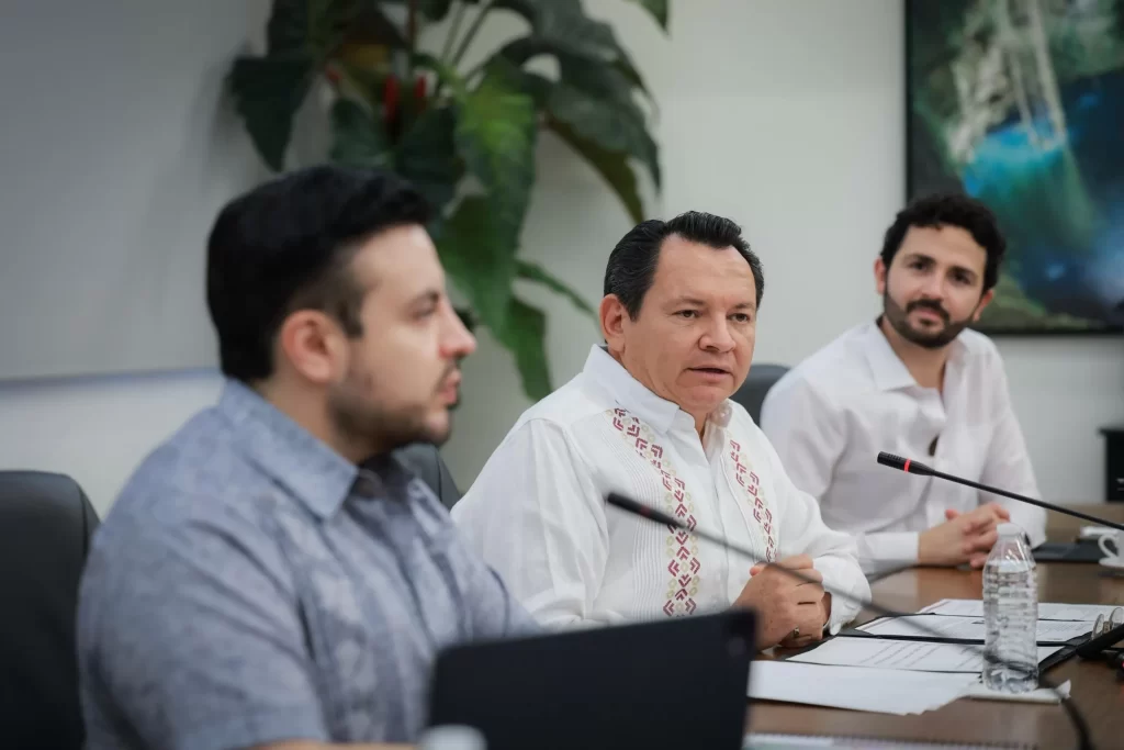 Tres hombres sentados en una mesa de juntas participando en una reunión, uno de ellos hablando mientras los otros escuchan