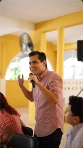 Hombre sonriente habla con micrófono frente a auditorio en lugar con paredes amarillas