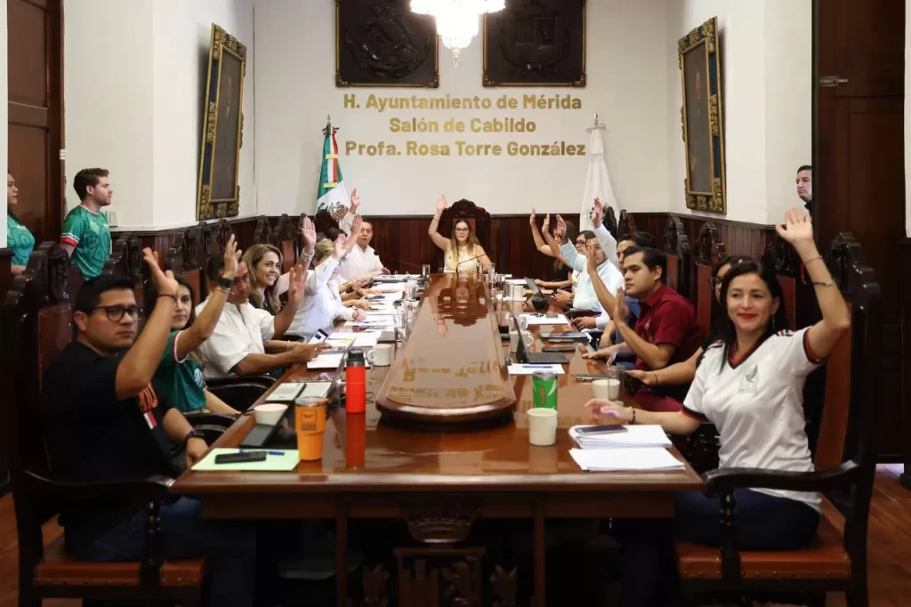 Personas en reunión formal dentro del Salón de Cabildo en Mérida levantando la mano durante votación