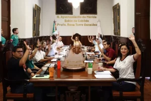 Personas en reunión formal dentro del Salón de Cabildo en Mérida levantando la mano durante votación