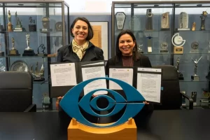 Dos mujeres sonrientes sostienen documentos firmados en sala con trofeos y escultura de ojo azul